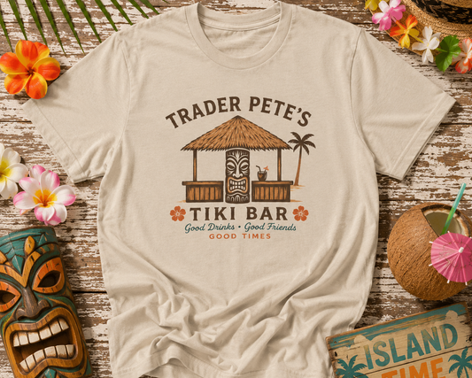 Beige t-shirt with 'Trader Pete's Tiki Bar' design on a wooden surface with tropical decorations.