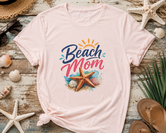 Pink t-shirt with 'Beach Mom' design on a wooden surface with beach-themed items.