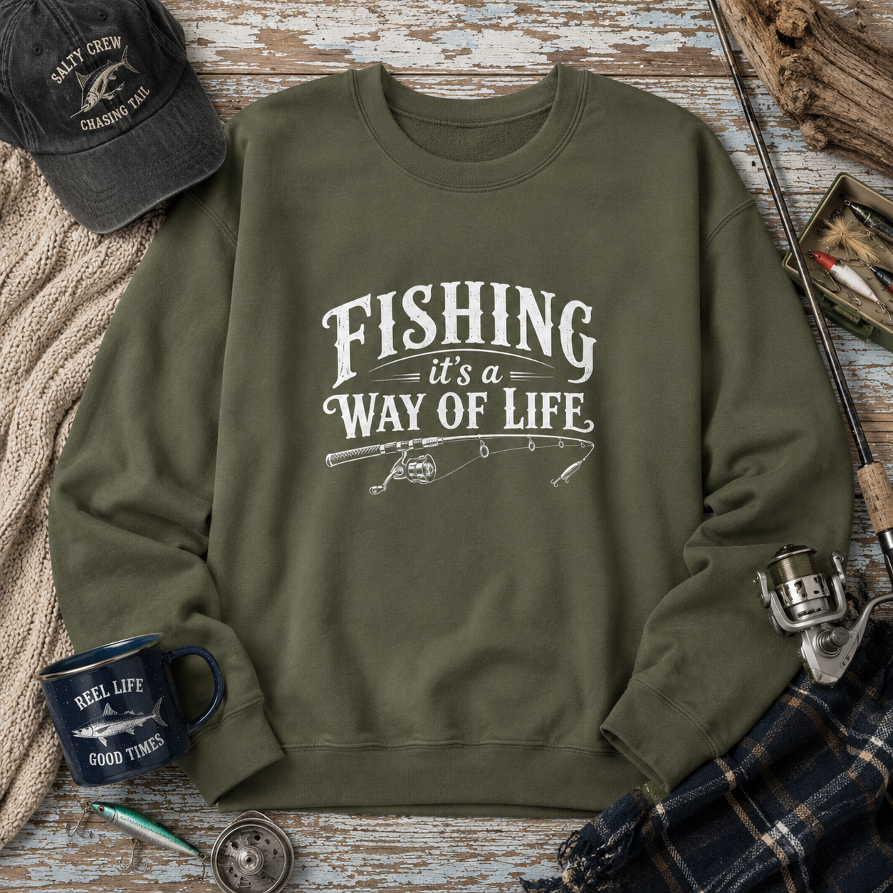 An olive green crewneck sweatshirt with a white graphic print reading 'FISHING it's a WAY OF LIFE' with a fishing rod illustration, styled in a flat lay on a rustic wooden background with a navy cap, mug, rod, and plaid fabric.