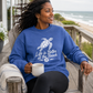 A woman sitting on a wooden beach porch wearing a blue crewneck sweatshirt with a white sea turtle graphic and the text 'Life is Better at the Beach'. She is holding a white mug, with the ocean and sandy beach visible in the background.