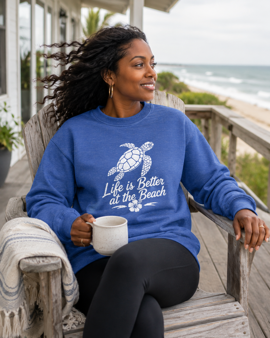 A woman sitting on a wooden beach porch wearing a blue crewneck sweatshirt with a white sea turtle graphic and the text 'Life is Better at the Beach'. She is holding a white mug, with the ocean and sandy beach visible in the background.