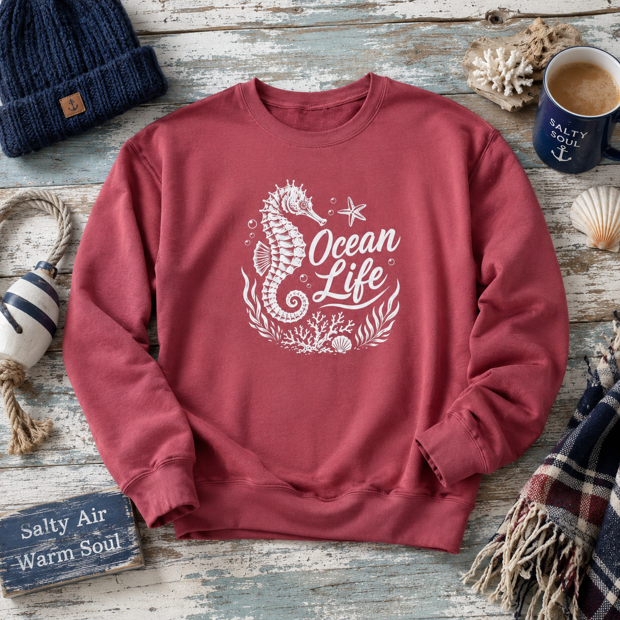 A dusty rose crewneck sweatshirt with a white 'Ocean Life' seahorse graphic, styled in a nautical flat-lay on a weathered wood background with a navy beanie, mug, and other coastal props.