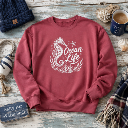A dusty rose crewneck sweatshirt with a white 'Ocean Life' seahorse graphic, styled in a nautical flat-lay on a weathered wood background with a navy beanie, mug, and other coastal props.