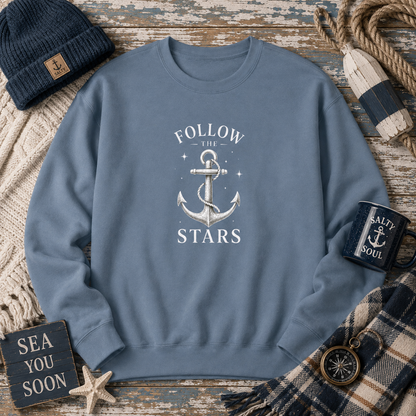A slate blue crewneck sweatshirt with a white anchor graphic and the text 'FOLLOW THE STARS' on the chest, styled in a nautical flat-lay with a beanie, rope, mug, scarf, and wooden sign.
