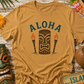 Beige t-shirt with 'Aloha' and tiki design on a wooden surface with tropical decorations.