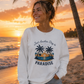 A woman stands on a beach at sunset wearing a white long-sleeve sweatshirt with a tropical graphic that reads 'Just Another Day in PARADISE' and denim shorts.