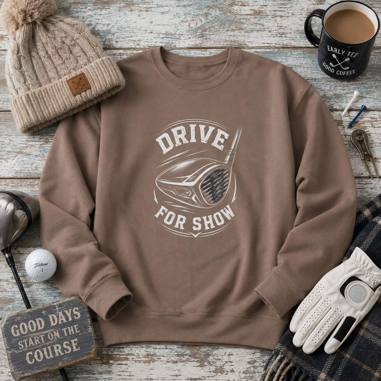 A brown crewneck sweatshirt with a white graphic reading 'DRIVE FOR SHOW' around a golf club head, styled in a flat lay on a weathered wood background with a beige knit beanie, coffee mug, golf club, glove, and other golf-themed props.