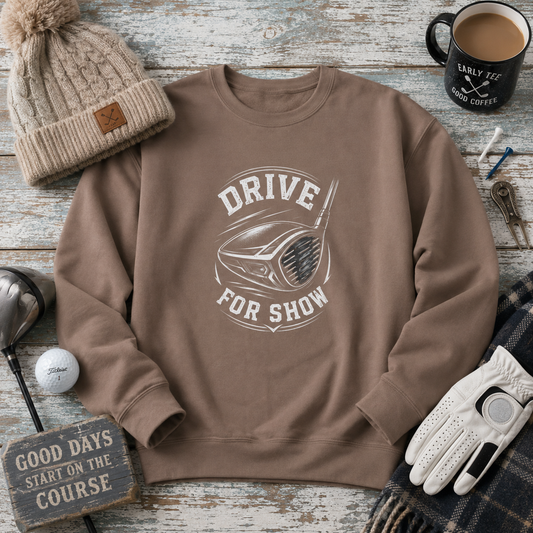 A brown crewneck sweatshirt with a white graphic reading 'DRIVE FOR SHOW' around a golf club head, styled in a flat lay on a weathered wood background with a beige knit beanie, coffee mug, golf club, glove, and other golf-themed props.