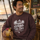 A man wearing a heathered dark plum crewneck sweatshirt with a white 'Tiki Bar' graphic, leaning on a wooden bar in a tropical beach setting.