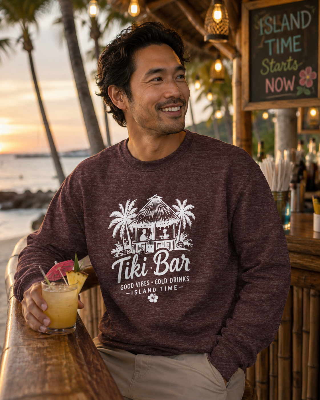A man wearing a heathered dark plum crewneck sweatshirt with a white 'Tiki Bar' graphic, leaning on a wooden bar in a tropical beach setting.
