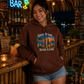 A woman wearing a brown hoodie with a colorful graphic that reads 'Great Minds Drink A Like' stands at a tiki bar counter. The hoodie features a sunset beach scene with palm trees.