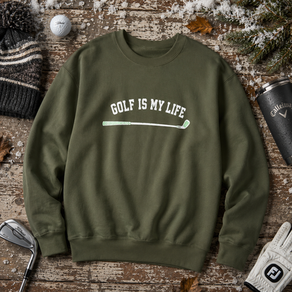 An olive green crewneck sweatshirt with the text 'GOLF IS MY LIFE' and a golf club graphic printed on the chest, styled in a flat lay on a rustic wooden surface with golf-themed props.