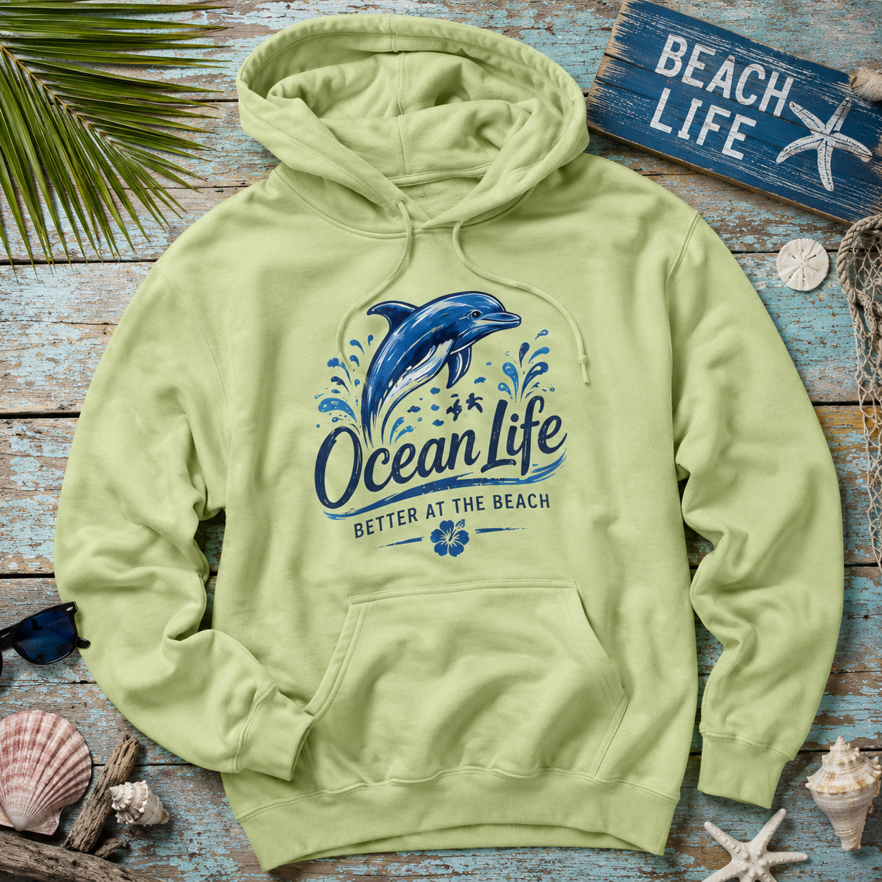 A light yellow-green pullover hoodie laid flat on a rustic blue wooden surface, surrounded by beach-themed props including shells, a starfish sign, sunglasses, and palm leaves. The hoodie features a large blue dolphin graphic with the words 'Ocean Life' and 'Better at the Beach' printed on the front.