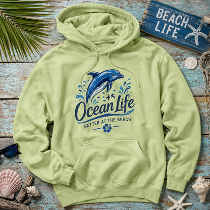 A light yellow-green pullover hoodie laid flat on a rustic blue wooden surface, surrounded by beach-themed props including shells, a starfish sign, sunglasses, and palm leaves. The hoodie features a large blue dolphin graphic with the words 'Ocean Life' and 'Better at the Beach' printed on the front.