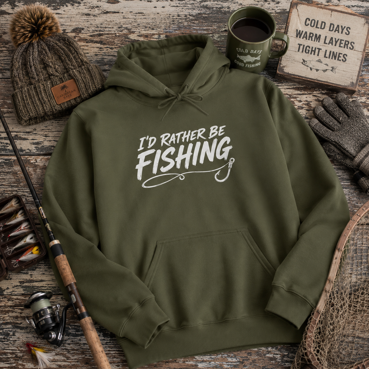 An olive green pullover hoodie with the white printed text 'I'D RATHER BE FISHING' and a fishing hook graphic, styled in a flat lay with a beanie, coffee mug, gloves, fishing rod, lures, and a rustic sign on a weathered wood background.