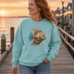 A woman with red hair stands on a wooden dock at sunset wearing a light aqua crewneck sweatshirt with a large colorful tropical fish graphic on the front, paired with blue jeans.