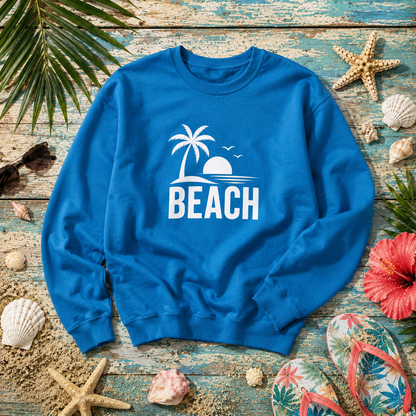 A bright blue crewneck sweatshirt with a white beach graphic and the word 'BEACH' printed on the front, styled in a flat lay on a weathered blue wooden surface with seashells, starfish, palm leaves, sunglasses, and floral flip-flops.