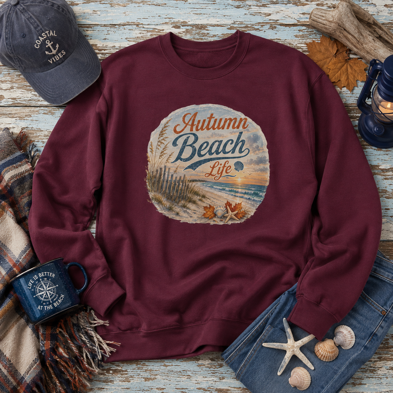 A maroon crewneck sweatshirt with a circular 'Autumn Beach Life' graphic on the chest, styled in a nautical flat-lay with a navy cap, denim shorts, a mug, and shells on a distressed wood background.