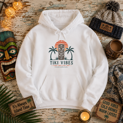 A white pullover hoodie with a 'Tiki Vibes' tropical sunset graphic, styled in a cozy flat-lay arrangement with a knit beanie, mug, candles, and rustic props on a weathered wood background.
