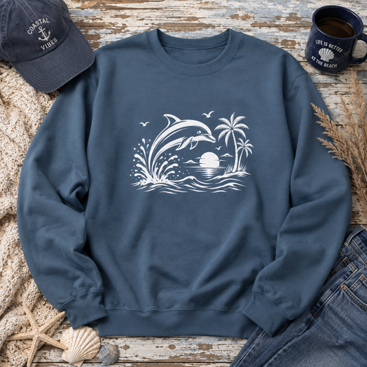 A navy blue crewneck sweatshirt with a white graphic of a dolphin, palm trees, and waves at sunset, styled in a beach-themed flat lay with a navy baseball cap, denim jeans, a mug, and seashells on a rustic wooden surface.