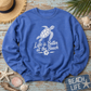 A heather blue crewneck sweatshirt with a white graphic of a sea turtle and the text 'Life is Better at the Beach' laid flat on a distressed blue wooden surface with beach-themed props including a straw hat, starfish, seashells, and a 'Beach Life' sign.