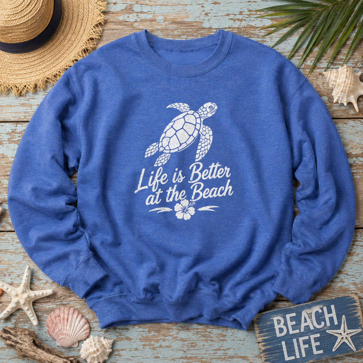 A heather blue crewneck sweatshirt with a white graphic of a sea turtle and the text 'Life is Better at the Beach' laid flat on a distressed blue wooden surface with beach-themed props including a straw hat, starfish, seashells, and a 'Beach Life' sign.