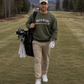a man walking in a fairway wearing  a Green sweatshirt with 'Golf Is My Life' printed on it against a white background