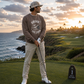A man stands on a golf course at sunset wearing a brown sweatshirt with a 'Drive for Show' graphic, khaki pants, and a baseball cap, holding a golf club. A small yard sign is visible on the grass next to him.