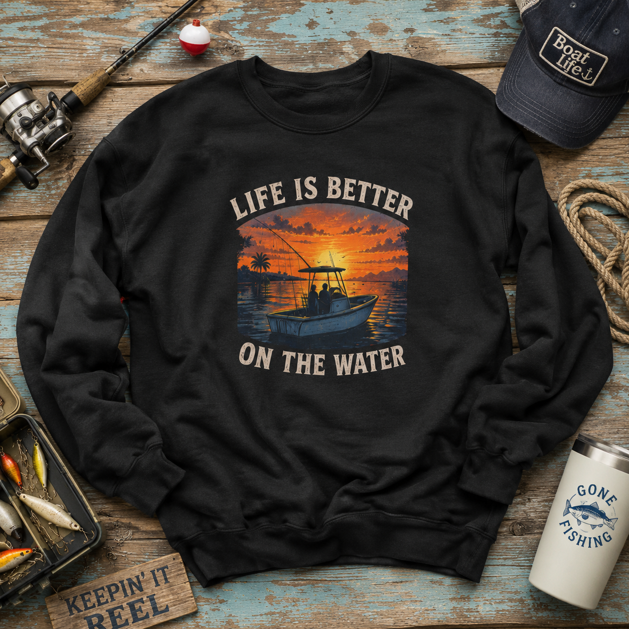 A black crewneck sweatshirt with a sunset fishing boat graphic and the text 'LIFE IS BETTER ON THE WATER' laid flat on a rustic blue wooden background with fishing-themed props.