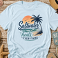 Light blue t-shirt with 'Saltwater heals everything' design on a wooden surface with beach-themed items.