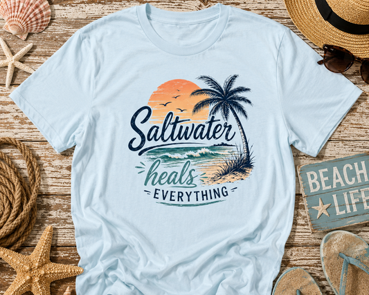 Light blue t-shirt with 'Saltwater heals everything' design on a wooden surface with beach-themed items.