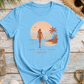 Light blue t-shirt with a beach-themed graphic on a wooden surface with beach accessories.